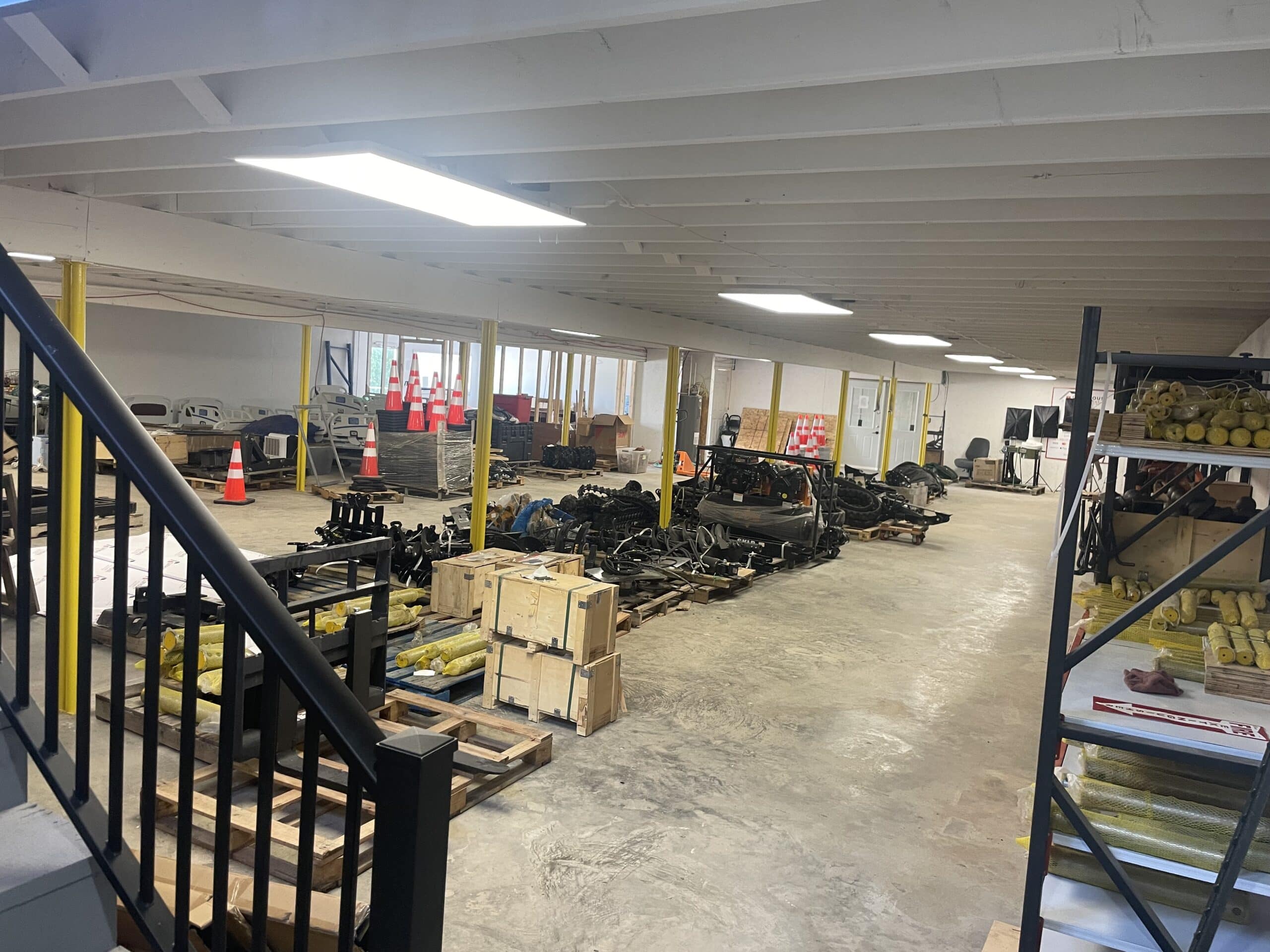 IMG_5648 - Cliffhouse Buildings Spacious warehouse interior with yellow support beams, stacks of equipment and attachments, and organized shelving along the walls.