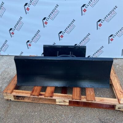 Mini skid steer snow pusher/dozer blade – wide black steel blade attachment with mounting plate resting on a wooden pallet in front of Cliffhouse Buildings-branded backdrop