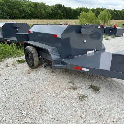 Long gray utility trailer with a built‑in metal storage box and diamond-plate fenders in a gravel lot.